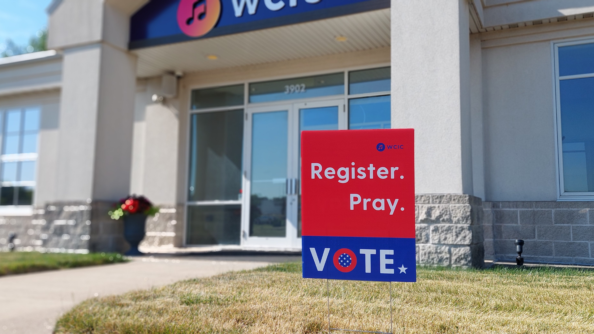 A picture of front of WCIC studios and a Yard sign that says Register Pray Vote